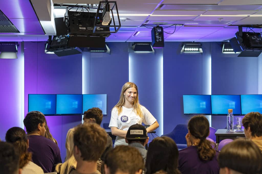 En ung kvinna i vit t-shirt med Rookie Startups logotyp på står framför en stor publik, inuti en studio med tv-skärmar.